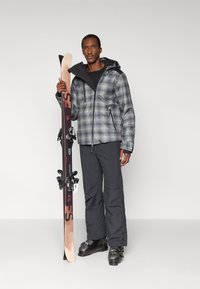 Conjunto de esquí con chaqueta gris de cuadros, capa base negra, pantalones grises y botas de esquí negras, sosteniendo un esquí con fijaciones negras y detalles de logo.