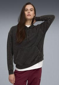 Frau in einem verblassten schwarzen Hoodie mit einer Fronttasche und bordeauxfarbenen Hosen, die mit einer Hand hinter dem Kopf vor einem grauen Hintergrund posiert.