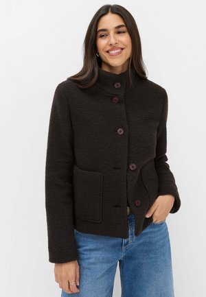 Kvinde iført en mørkebrun knappet jakke med høj krave og forlommer, kombineret med blå jeans, smilende og kigger sidelæns.
