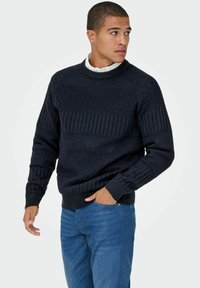 Pull en tricot navy avec des motifs en diamant texturés et des côtes. Encolure ras du cou et manches longues. Assorti avec un jean bleu.