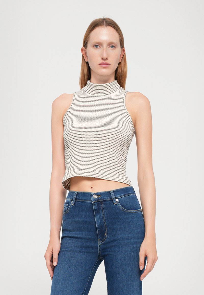 Vrouw in een mouwloze, hooggesloten gestreepte crop top en hoge taille blauwe jeans, staande tegen een effen witte achtergrond.