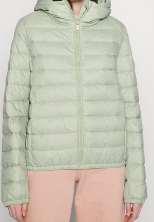 Persoon met lichtgroene gewatteerde pufferjas met capuchon en ritssluiting aan de voorkant, gecombineerd met lichtroze broek, tegen een effen achtergrond.
