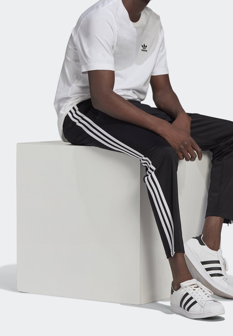 T-shirt bianca in cotone con logo nero, pantaloni sportivi neri con strisce laterali bianche e sneaker bianco e nere, seduto su un cubo bianco.