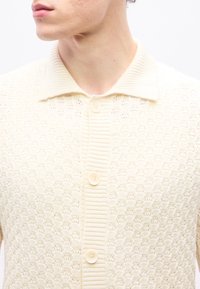 Homme portant un cardigan en tricot de couleur crème avec un motif texturé, un col côtelé et trois boutons à l'avant.