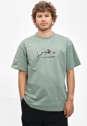 Junger Mann mit lockigem Haar, der ein grünes T-Shirt mit einem Cartoon-Enten- und Schwimmer-Motiv auf der Brust, schwarze Hose und ein blaues Armband trägt.