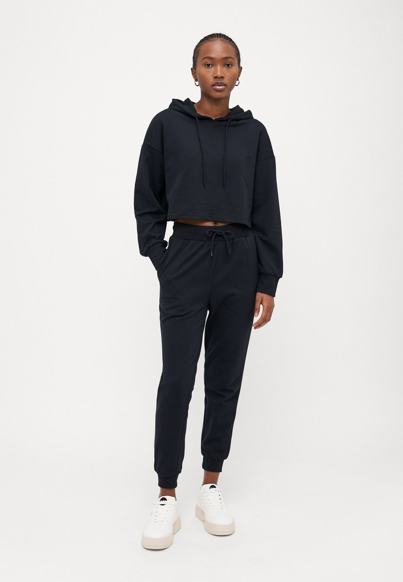 Felpa nera corta con cappuccio, coulisse e maniche lunghe, abbinata a pantaloni jogger coordinati con vita elastica e polsini.