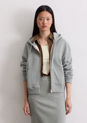 Haut de sweat à capuche zippé gris en tissu doux, avec une poche avant, doté d'une capuche avec cordon de serrage. Porté par-dessus une chemise à carreaux et une jupe ajustée.