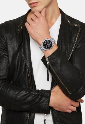 Montre-bracelet en acier inoxydable argenté avec un cadran noir, trois sous-cadrans, et une veste en cuir noir portée sur un t-shirt blanc.