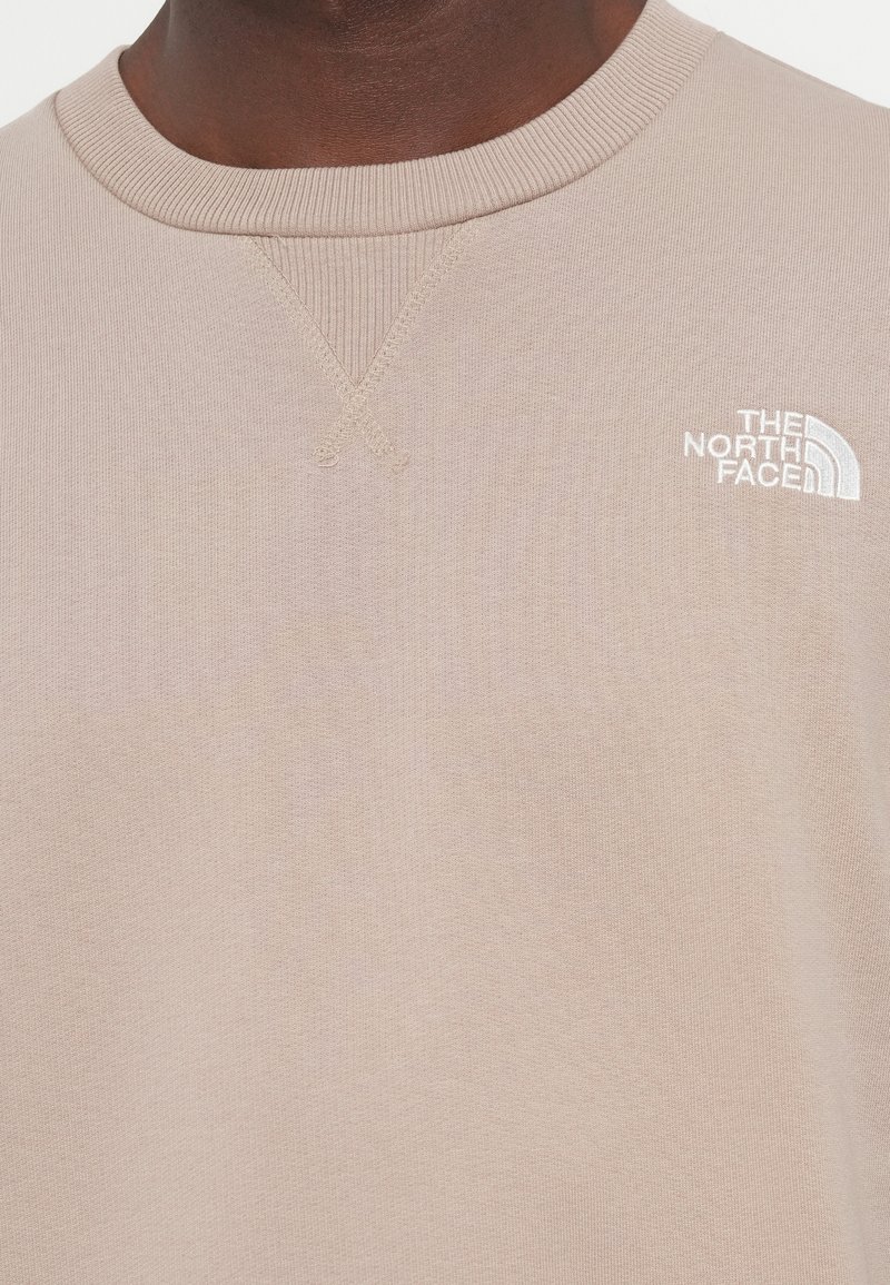 Beige Sweatshirt mit geripptem Rundhalsausschnitt und subtilen Kreuzstichdetails. Mit einem weißen bestickten Logo auf der Brust. Weicher Stoff.
