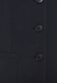 Blazer negro con una textura suave, cierre de botones y un bolsillo lateral. Incluye cuatro botones negros con cuatro orificios cada uno.