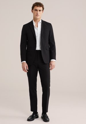 Giovane uomo in piedi, indossa un abito nero con camicia bianca e mocassini neri su uno sfondo chiaro uniforme.