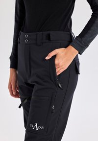 Zwarte skibroek met een gladde textuur, voorzien van een knoopsluiting, meerdere ritszakken en een logo op het onderbeen.