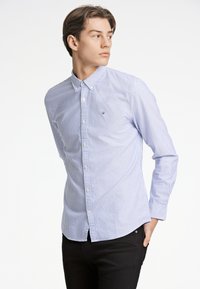 Camicia button-up in cotone leggero, con righe verticali blu e bianche. Include colletto, maniche lunghe e un piccolo logo sul petto.