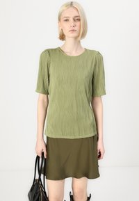 Top de manga curta, verde-oliva, com padrão texturizado em ondas; decote redondo; combinado com uma saia simples verde-oliva; modelo segura uma bolsa preta.