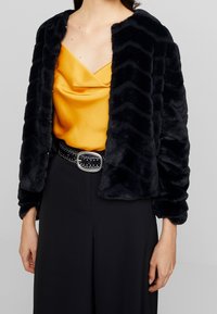 Femme portant une veste en fausse fourrure noire sur un haut en satin jaune moutarde, associée à un pantalon noir et une ceinture noire cloutée avec une boucle argentée.