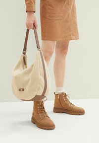 Beżowa torebka z futrzanym wykończeniem i brązowymi skórzanymi detalami oraz paskiem, w połączeniu z beżowymi butami na sznurówki. Tło ma jasny, neutralny kolor.