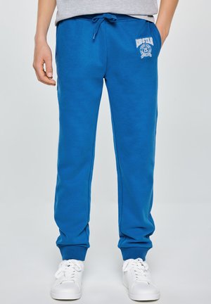 Mężczyzna noszący królewsko niebieskie spodnie dresowe z białym logo na lewym udzie, w połączeniu z białymi sneakersami i szarą koszulą schowaną w spodnie.