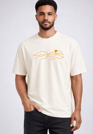 Man with short curly hair and beard wearing a cream t-shirt with an orange mountain and sun line drawing, standing with one hand in pocket.