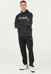 Svart huvtröja med vitt "FZ FORZA"-logotyp, kombinerad med svarta träningsbyxor. Modellen har på sig vita träningsskor. Enkel, avslappnad sportig stil.