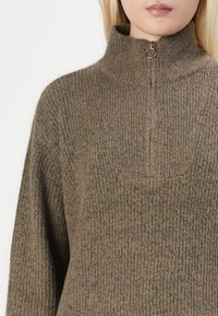 Maglione marrone a coste con colletto alto, dotato di una chiusura a metà cerniera e tessuto strutturato. Il design presenta un motivo sottile per un tocco di dettaglio in più.