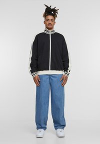 Sort zip-up jakke med hvide stribede detaljer, parret med løse lyseblå jeans og hvide sneakers. Har høj krave og sidelommer.