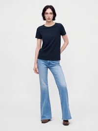 Mujer con el pelo corto y oscuro que lleva una camiseta negra lisa, vaqueros acampanados azul claro y zapatos marrones, de pie frente a un fondo blanco.