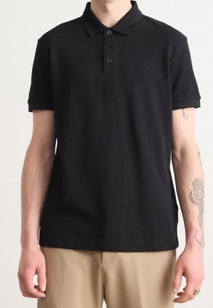 Polo noir en coton doux. Comprend un col, une patte de boutonnage à trois boutons et un logo brodé. Porté avec un pantalon beige clair.