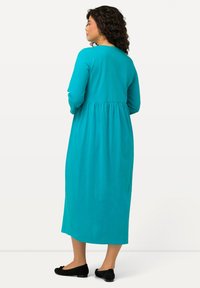 Turkusowa sukienka z długim rękawem, z zebranym w pasie krojem, zwiewnym dołem i odkrytymi plecami. Noszona z czarnymi balerinami, cechującymi się gładką fakturą materiału.