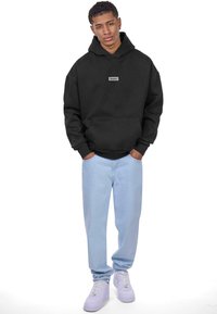 Sweatshirt à capuche noire avec une poche avant et un écusson logo bleu clair, assorti à un jean bleu clair et des baskets blanches.
