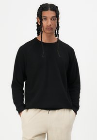 Sweatshirt noir à manches longues en tissu texturé avec des poignets côtelés, arborant une coupe décontractée et un col rond, associé à un pantalon de couleur claire.