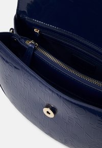 Námořnická modrá lakovaná kůže kabelka s embosovaným textem "NOVA VALENTINI", zlatý zip a magnetické zapínání částečně otevřené, ukazující vnitřní kapsy.