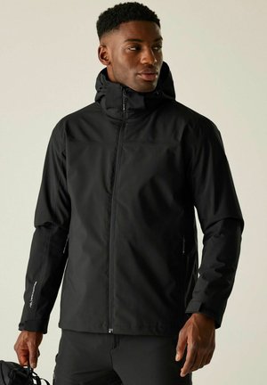 Homme portant une veste à capuche noire avec des poches zippées et tenant un sac noir, regardant sur le côté sur un fond uni.