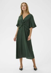 Rochie verde înfășurată, cu decolteu adânc în V, mâneci flutter și detaliu noduros în față. Material neted, până la genunchi, asortată cu tocuri negre ascuțite.