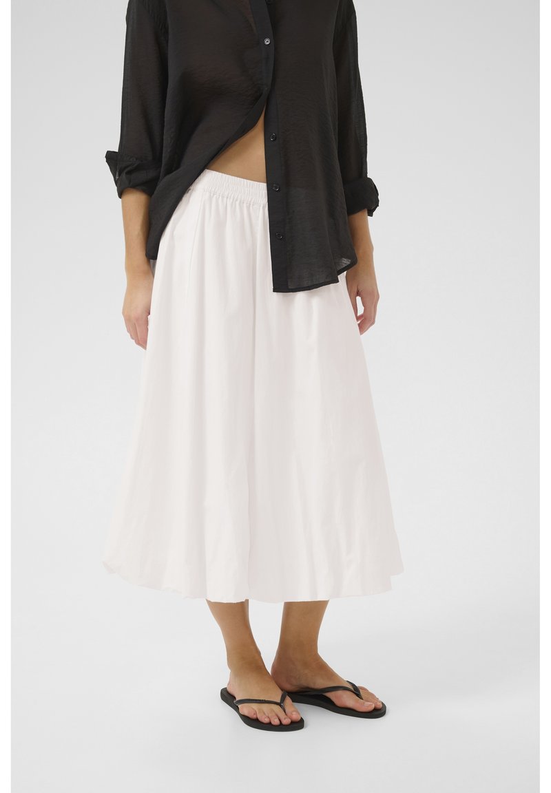 Jupe blanche, longueur genou, en tissu doux et léger avec une taille élastique. Associée à une chemise noire transparente à boutons et des tongs.