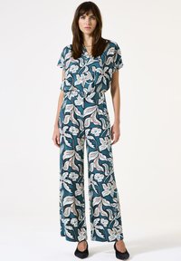 Blumenmuster-Jumpsuit in Teal und Creme, mit kurzen Ärmeln, einem Band in der Taille, weitem Bein und schwarzen Ballerinas. Weicher Stoff mit strukturiertem Design.