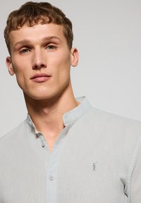 Ljusgrå skjorta med krage, knäppning, texturerad tyg och en liten logotyp broderad i marinblått på vänster sida.