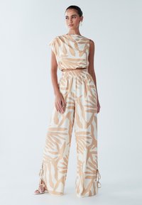 Bēša un baltas divdaļīgas tērps ar īsu topu, kam ir asimetrisks kakla izgriezums, un plašām biksēm ar abstraktiem rakstiem.