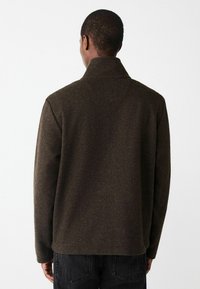 Hnedý sveter z vlnenej zmesi s vysokým golierom, voľným strihom a dlhými rukávmi. Textúrovaná tkanina má jemný pocit, poskytuje teplo a pohodlie.