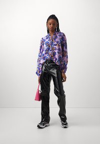 Blusa floral púrpura con mangas largas, pantalones negros brillantes, zapatillas blancas y negras, y un pequeño bolso rosa.
