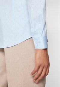 Světle modrá košile na knoflíky s jemnou vzorovanou strukturou, dlouhými rukávy a detailem na manžetách, v kombinaci s béžovými látkovými kalhotami.