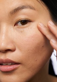 Nahaufnahme eines Gesichts, auf dem eine Person eine leichte Creme unter dem Auge aufträgt, die eine glatte, leicht glänzende Hauttextur zeigt.