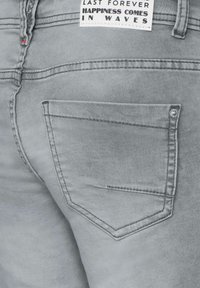 Jeans in denim grigio con una singola tasca posteriore caratterizzata da un dettaglio di cucitura orizzontale e un rivetto metallico, etichettati con un'etichetta bianca in vita.