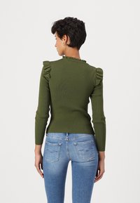 Geribbelde, lange mouwen top in olijfgroen met geschulpte schouderaccenten en een hoge kraag; gecombineerd met aansluitende blauwe jeans.