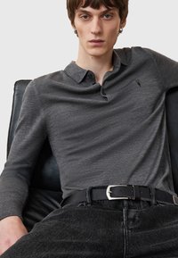 Grå langærmet polo med krave og tre knapper, med et diskret logo. Kombineret med sorte jeans og et sort bælte med sølvspænde.