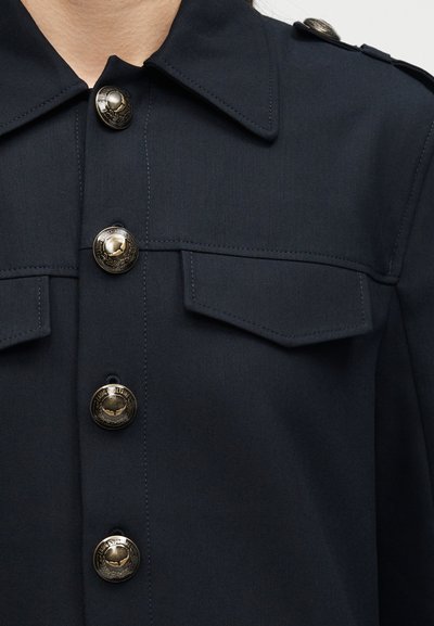 Μπλε ναυτικό μπουφάν με μυτερό γιακά, διακοσμητικά μεταλλικά κουμπιά με ανάγλυφο κείμενο, πλαϊνές τσέπες στο στήθος με καπάκι και επωμίδες στους ώμους.