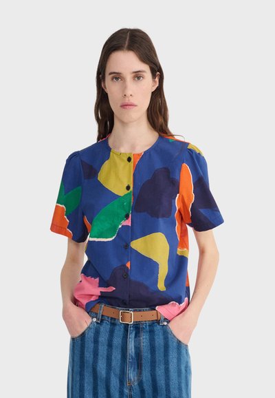 Femme portant une chemise boutonnée avec des formes abstraites multicolores en bleu, vert, jaune, orange et rose, associée à un jean rayé bleu.