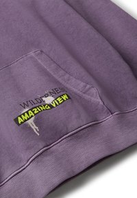 Lila Stoff mit einer Fronttasche, die schwarzen, weißen und neon-gelben Text mit der Aufschrift "Wilderness Amazing View" zeigt.