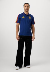 Jūras spēku polos krekls ar sarkaniem un dzelteniem svītrainiem piedurkņu daļās, izšūts ģerbonis, saskaņots ar melniem platām biksēm un baltiem sneakeriem.