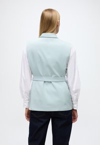 Vestă albastru deschis cu un design croită, cu curea în talie, purtată peste o cămașă albă cu mânecă lungă, combinată cu blugi albastru închis.