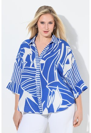 Femme blonde portant une chemise boutonnée à manches larges avec un motif abstrait bleu et blanc, et un pantalon blanc, debout devant un fond clair.
